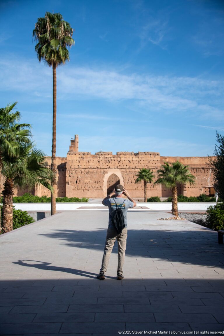 Visitor bending forward to take a photograph across the palm-lined central court of Badii Palace.