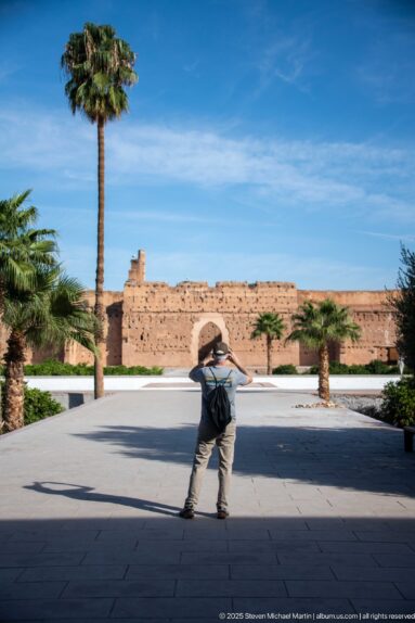 Visitor bending forward to take a photograph across the palm-lined central court of Badii Palace.