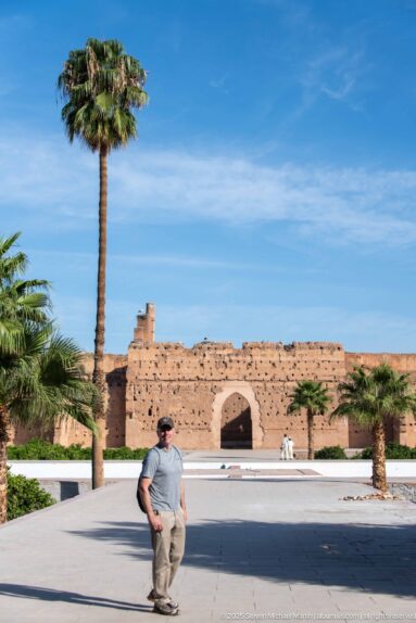 Visitor walking across the broad central court of Badii Palace with palms and a long earthen wall behind him.