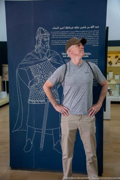 Visitor posing in front of a large dark exhibition panel with a line drawing of an armed historical figure.