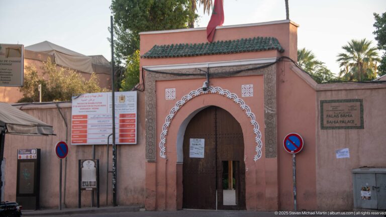 Salmon-pink Bahia Palace entrance with green-tiled canopy, Moroccan flag, arched wooden doorway and official signs in Marrakech.