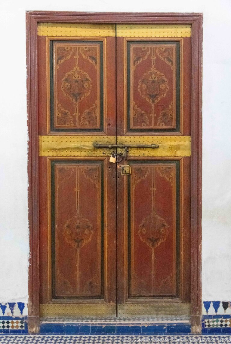 Closed reddish wooden double doors with painted floral motifs, brass-colored bands and latch at Bahia Palace.