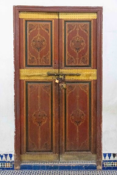 Closed reddish wooden double doors with painted floral motifs, brass-colored bands and latch at Bahia Palace.