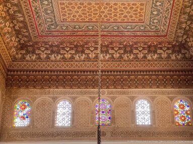 Painted Bahia Palace ceiling above a row of small arched white and multicolored stained-glass windows.