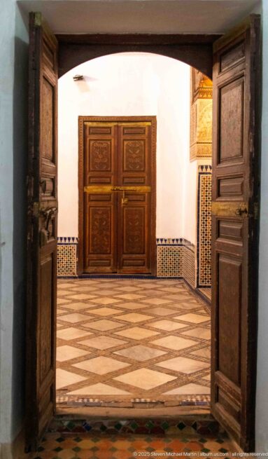 Open wooden doors framing a Bahia Palace room with diamond-pattern floor, zellij dado and closed double doors opposite.