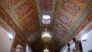 Long polychrome Bahia Palace ceiling with two chandeliers and a small raised central roof opening.