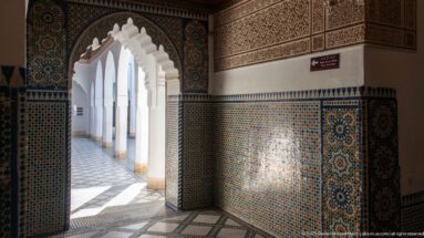 Zellij-lined Bahia Palace doorway opening onto a sunlit white arcade with repeating scalloped arches.