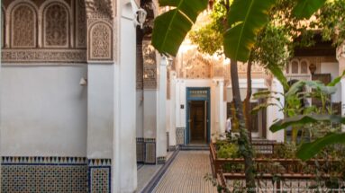 Zellij-lined passage through a planted Bahia Palace courtyard, ending at a turquoise door beneath carved and painted decoration.