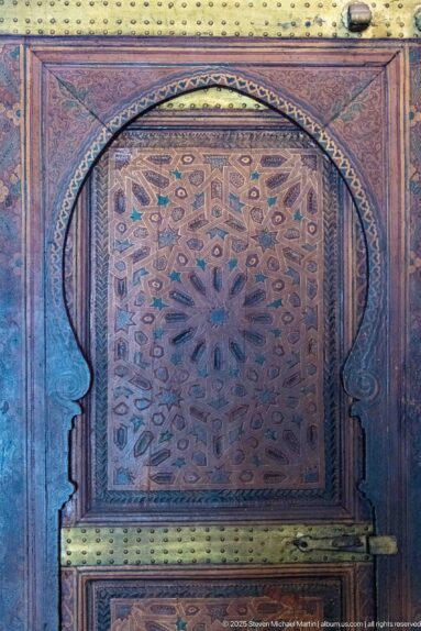 Carved Bahia Palace wooden door with radiating geometric star panel and broad brass-colored horizontal strap.