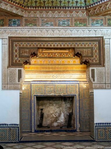 Large Bahia Palace fireplace with dark hearth, geometric zellij, carved plaster panels and projecting golden-toned hood.