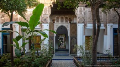 Ornamented Bahia Palace courtyard façade with carved plaster, zellij, central doorway and banana plants in the foreground.