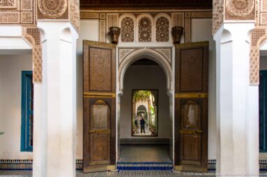Open carved wooden doors beneath an ornate arch, with patterned tile floor and a view into a garden court.