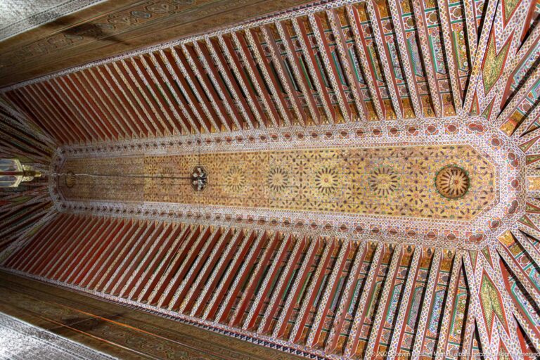 Symmetrical upward view of a richly painted Bahia Palace timber ceiling with colorful beams and floral-geometric central decoration.