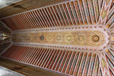 Symmetrical upward view of a richly painted Bahia Palace timber ceiling with colorful beams and floral-geometric central decoration.
