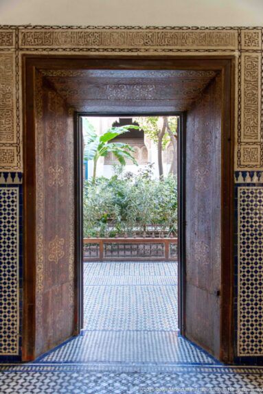 Open wooden doorway at Bahia Palace framing a leafy courtyard across patterned zellij flooring.