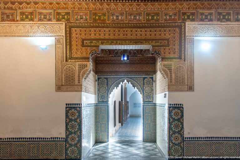 Frontal Bahia Palace doorway with zellij tiles, carved plaster, painted wood and a long passage visible beyond.