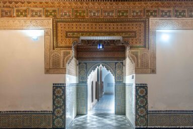 Frontal Bahia Palace doorway with zellij tiles, carved plaster, painted wood and a long passage visible beyond.