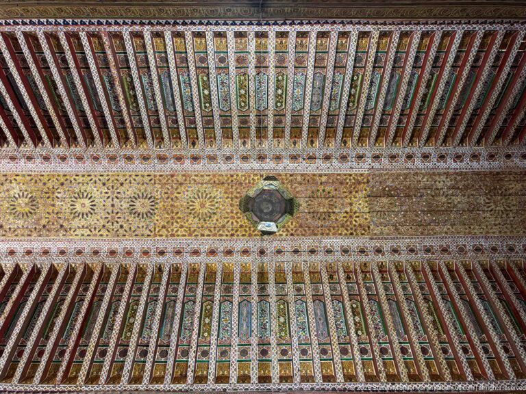 Symmetrical painted timber ceiling at Bahia Palace with repeated colored beams, ornate central strip and hanging light.