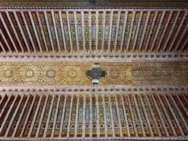 Symmetrical painted timber ceiling at Bahia Palace with repeated colored beams, ornate central strip and hanging light.