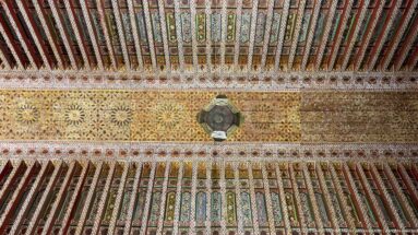 Polychrome Bahia Palace timber ceiling with repeated rosettes, multicolored beams and a central hanging fixture.
