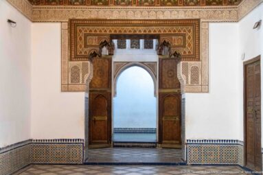 Open wooden doorway at Bahia Palace framed by zellij and carved plaster beneath a projecting painted timber lintel.