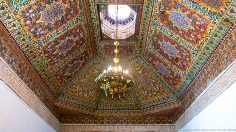 Ornate polychrome coffered ceiling at Bahia Palace with floral-geometric panels, central raised opening and hanging chandelier.