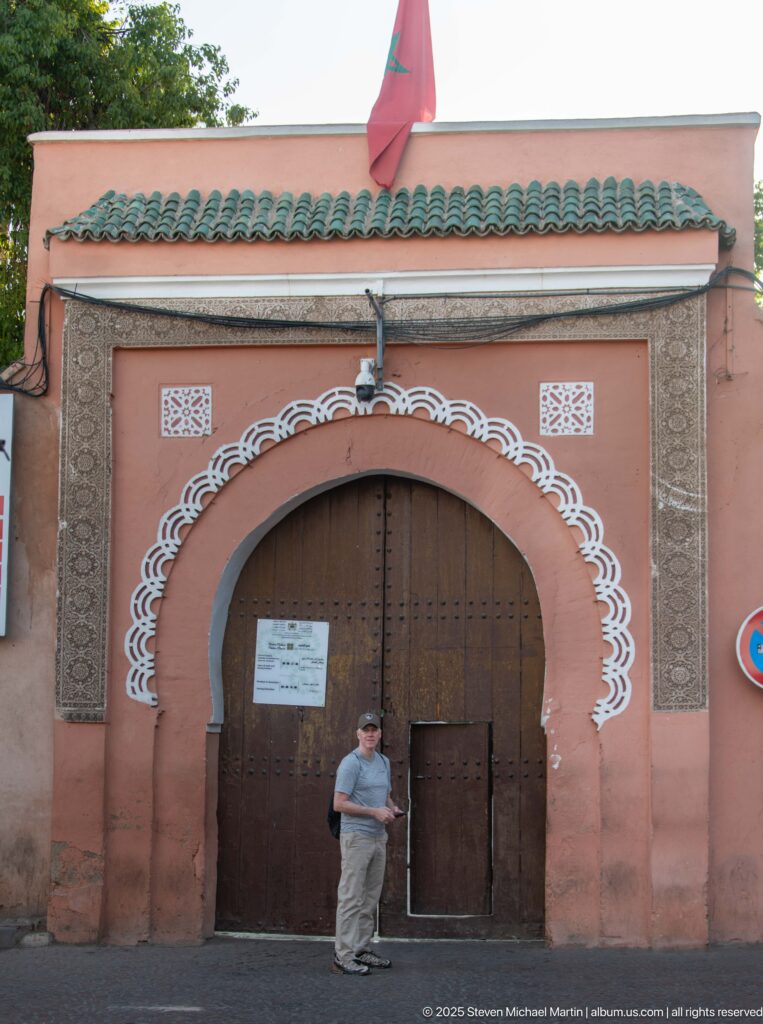Visitor standing before Bahia Palace’s salmon-pink entrance, with studded wooden doors, white arch ornament, green tiles and Moroccan flag.