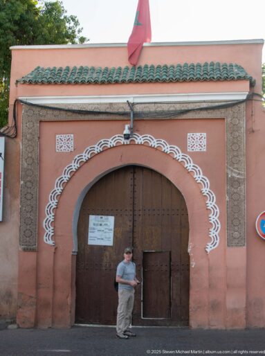 Visitor standing before Bahia Palace’s salmon-pink entrance, with studded wooden doors, white arch ornament, green tiles and Moroccan flag.