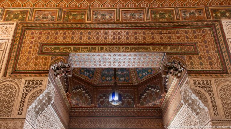 Upward view of a richly decorated Bahia Palace doorway with painted wood, carved plaster, calligraphic bands and a hanging lantern.