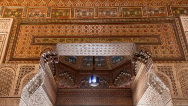 Upward view of a richly decorated Bahia Palace doorway with painted wood, carved plaster, calligraphic bands and a hanging lantern.