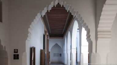 Long Bahia Palace arcade with white scalloped arches, dark timber ceiling, open wooden doors and bright side openings.