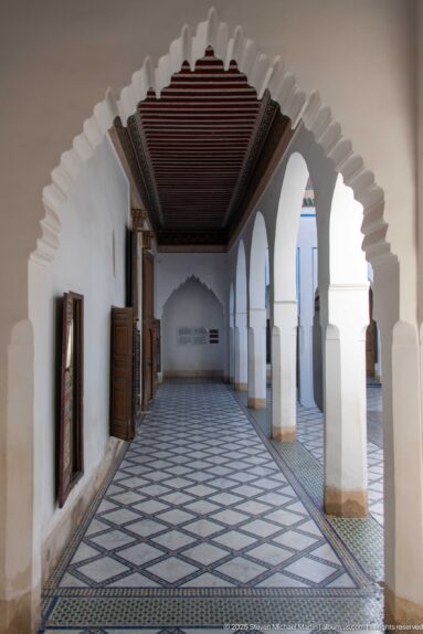 Long Bahia Palace gallery with white scalloped arches, dark timber ceiling, open shutters and geometric tiled floor.
