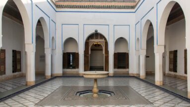 Symmetrical Bahia Palace courtyard with white arcades, patterned paving and a small pedestal fountain at center.