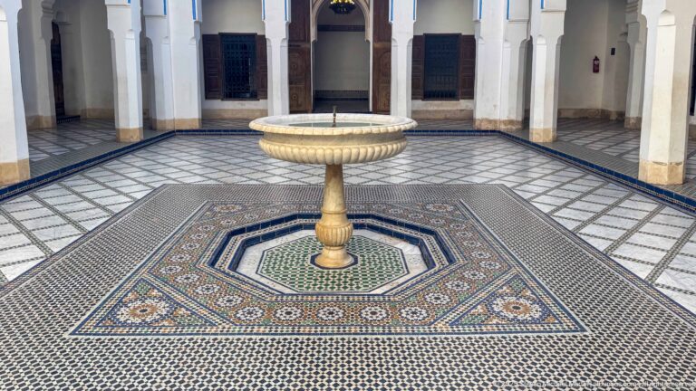 Stone pedestal fountain centered within multicolored geometric zellij paving in a Bahia Palace courtyard.