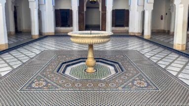 Stone pedestal fountain centered within multicolored geometric zellij paving in a Bahia Palace courtyard.