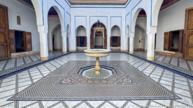 Wide Bahia Palace courtyard with central fountain, geometric tiled floor, white arcades and open wooden doors.