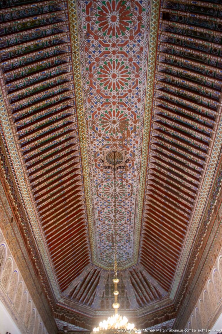 Long Bahia Palace timber ceiling with floral central panel, dark red beams and hanging light.