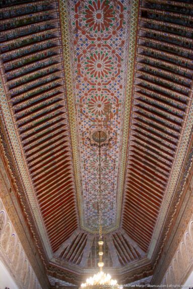 Long Bahia Palace timber ceiling with floral central panel, dark red beams and hanging light.