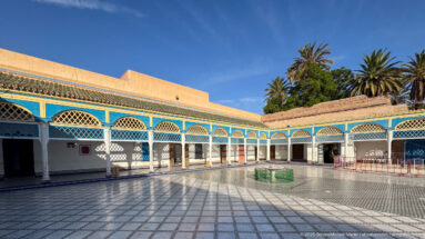 Large Bahia Palace courtyard with blue-and-white arcades, geometric paving, central green-edged basin and palms beyond.