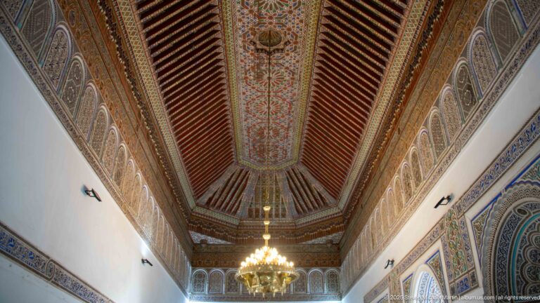 Long Bahia Palace reception room ceiling with painted central panel, dark beams, chandelier and repeated wall lunettes.