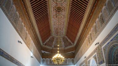 Long Bahia Palace reception room ceiling with painted central panel, dark beams, chandelier and repeated wall lunettes.