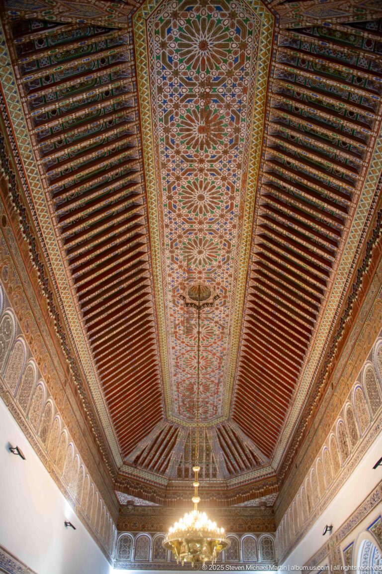 Symmetrical Bahia Palace ceiling with floral central strip, reddish timber beams and the edge of a chandelier below.