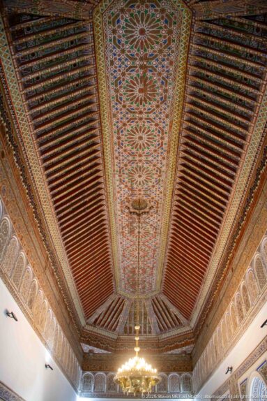 Symmetrical Bahia Palace ceiling with floral central strip, reddish timber beams and the edge of a chandelier below.