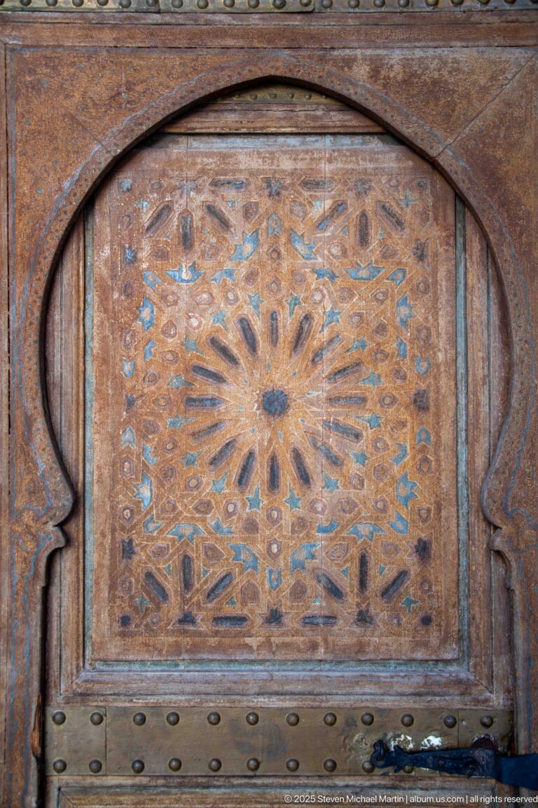 Weathered Bahia Palace wooden door panel with carved radial medallion, pointed arch frame and metal studs.