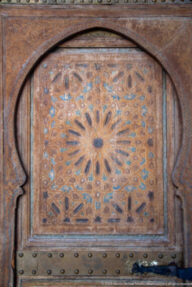 Weathered Bahia Palace wooden door panel with carved radial medallion, pointed arch frame and metal studs.