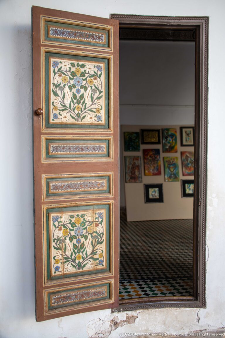 Single open floral-painted wooden door at Bahia Palace leading to a tiled room with framed artworks on the wall.