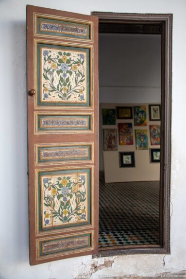 Single open floral-painted wooden door at Bahia Palace leading to a tiled room with framed artworks on the wall.