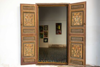 Open floral-painted double doors at Bahia Palace framing a tiled room with framed works on white walls.