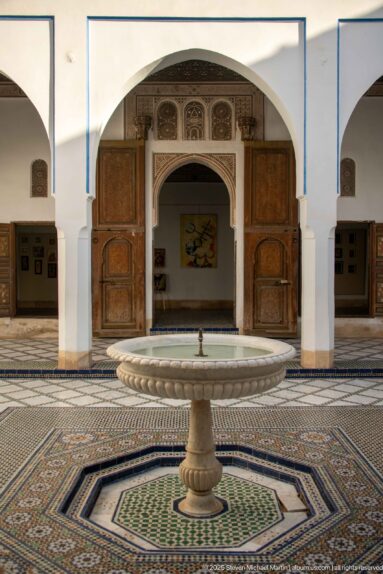 Bahia Palace pedestal fountain centered before an arched doorway, open wooden doors and geometric zellij paving.