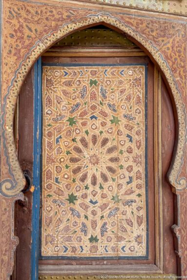 Painted geometric star panel in ochre, green, red and blue set within a horseshoe-shaped wooden frame.